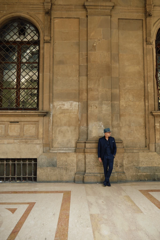Style by Sarai editorial — man in navy three-piece pinstripe suit and patterned fedora against a Florentine stone loggia during Pitti Immagine Uomo 107.