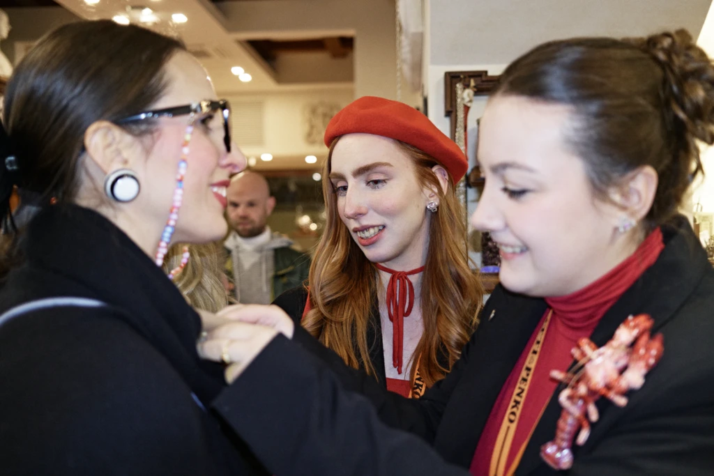 Evening in the streets of Florence during Pitti Immagine Uomo 107 — three women chatting inside a jewellery shop, one wearing a red beret and a coral lobster brooch.