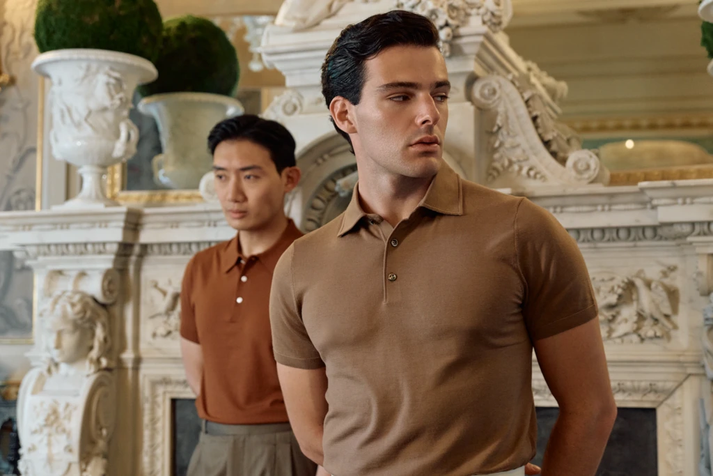 Common Suites fashion editorial at Villa Cora, Florence: two models — one in a caramel knit polo, one in a rust orange knit polo — standing side by side in front of the villa's hand-carved white marble fireplace.