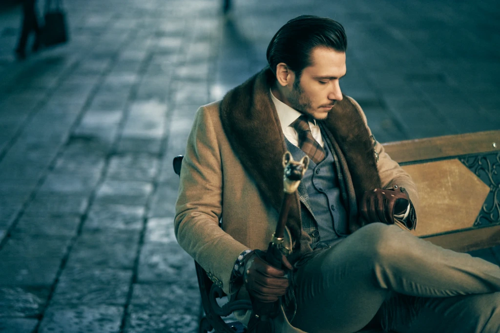 Elegant dandy menswear portrait in Florence: model in a camel wool coat with fur collar, grey tweed waistcoat, tartan tie and brown leather gloves, seated on an ornate bench holding a French bulldog cane on wet cobblestone.