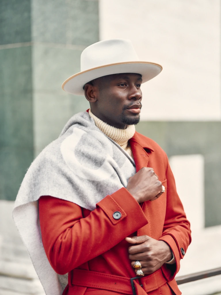 Contemporary menswear portrait: stylish man in a rust-red wool overcoat, cream ribbed turtleneck and off-white wide-brim fedora, with a grey and white patterned scarf against a soft grey wall.