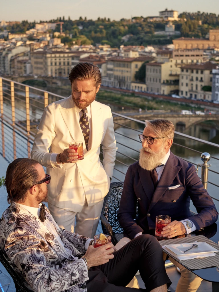 Florence rooftop lifestyle scene: three gentlemen in sartorial white and navy tailoring enjoying Negroni cocktails at golden hour with a panoramic view over the Arno river and the Oltrarno hills.