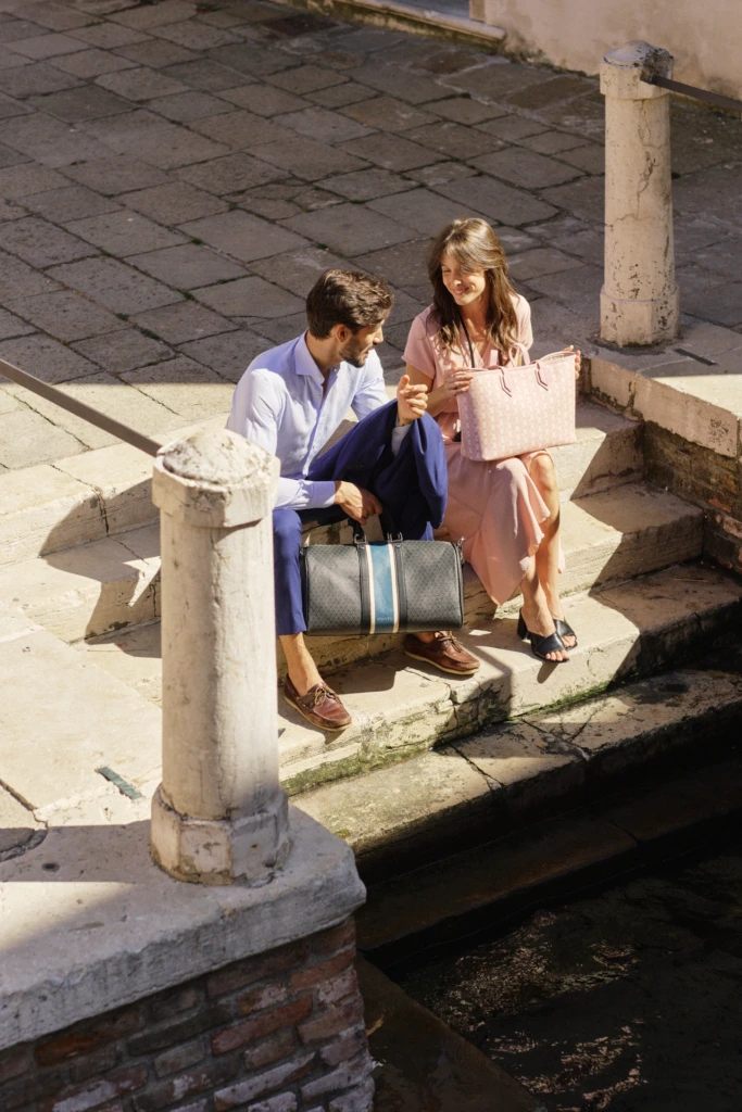 Fashion travel lifestyle in Venice: young couple sitting on stone canal steps with Vonto luxury duffel and tote bags, styled in a soft blue linen shirt and a pink summer dress.