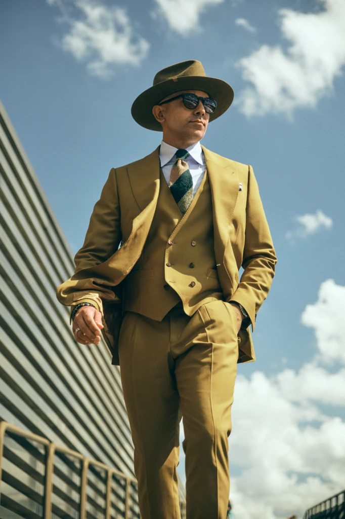 Pitti Uomo street style: low-angle portrait of a bespoke enthusiast in a mustard three-piece suit with striped shirt, patterned tie, brown wool fedora and sunglasses against a bright sky.