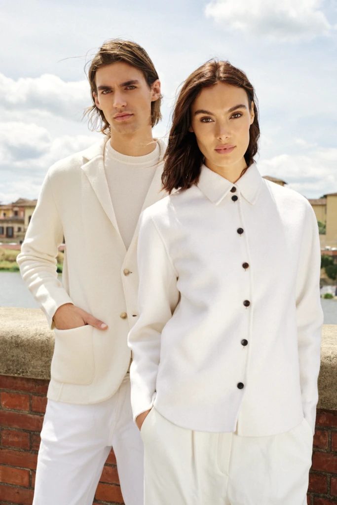 Minimal couple portrait in Florence on the Arno: a man in cream cashmere blazer and a woman in a white shirt-jacket, styled in coordinated ivory tailoring on a brick parapet.