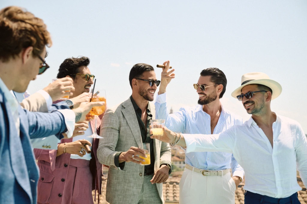 Pitti Uomo rooftop lifestyle scene in Florence: five stylish men in linen suits, striped shirts and Panama hats sharing a toast with Aperol Spritz cocktails and a cigar under a bright summer sky.