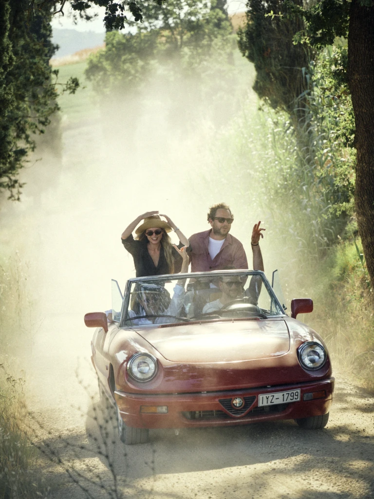 Vintage Tuscan road-trip lifestyle photograph: friends laughing and waving from a red Alfa Romeo Spider convertible with Italian plates, driving down a dusty cypress-lined country lane.