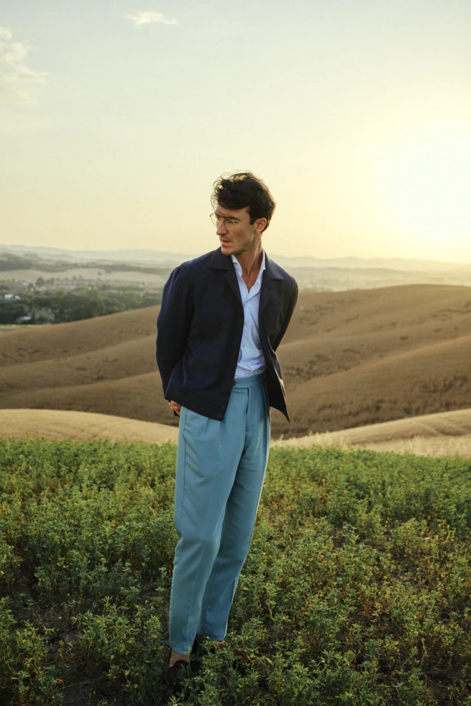 Fashion menswear editorial in the Tuscan hills at sunset: man in a navy overshirt, white shirt and light-blue high-waisted trousers with round glasses, standing between rolling wheat fields.