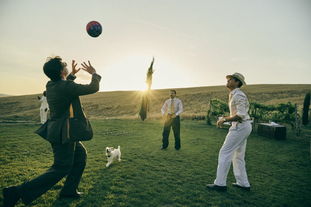 Playful Tuscan lifestyle scene at sunset: group of gentlemen in tailored suits playing with a decorative ball and a white West Highland terrier on a lawn beside a vineyard and cypress tree.
