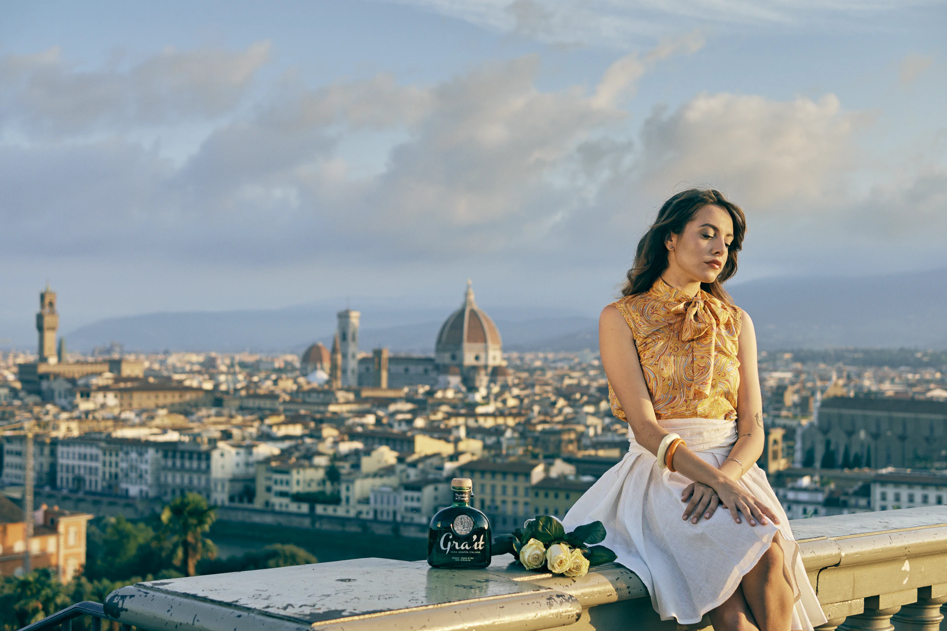 Model in a gold-patterned blouse with the Gra'it grappa bottle and white roses, overlooking the Florence skyline at sunset