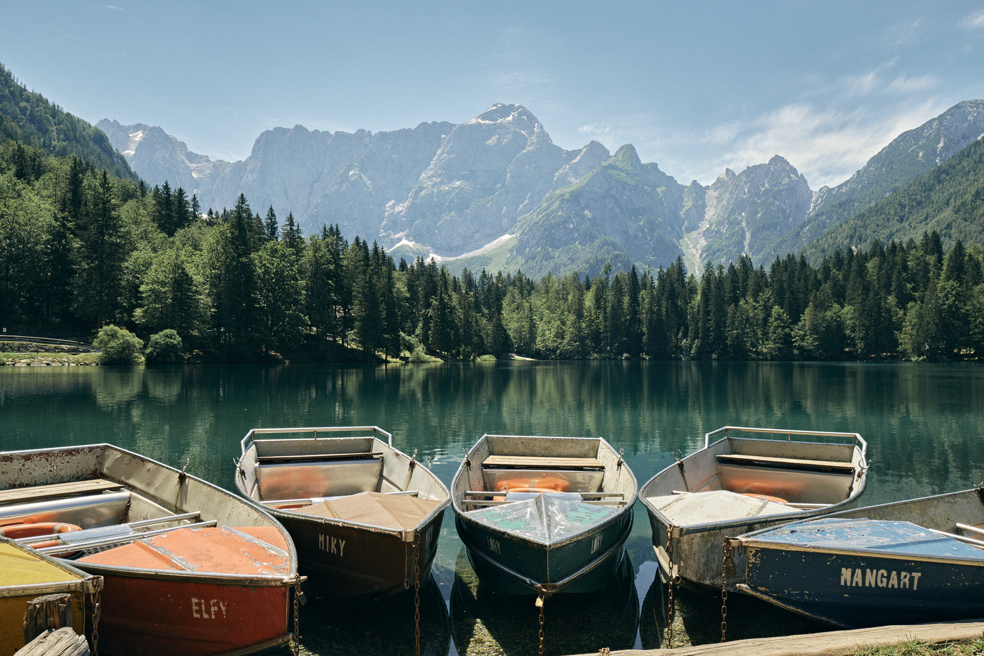 Wooden rowboats named Elfy, Miky and Mangart moored on emerald Fusine Lake beneath the jagged Julian Alps