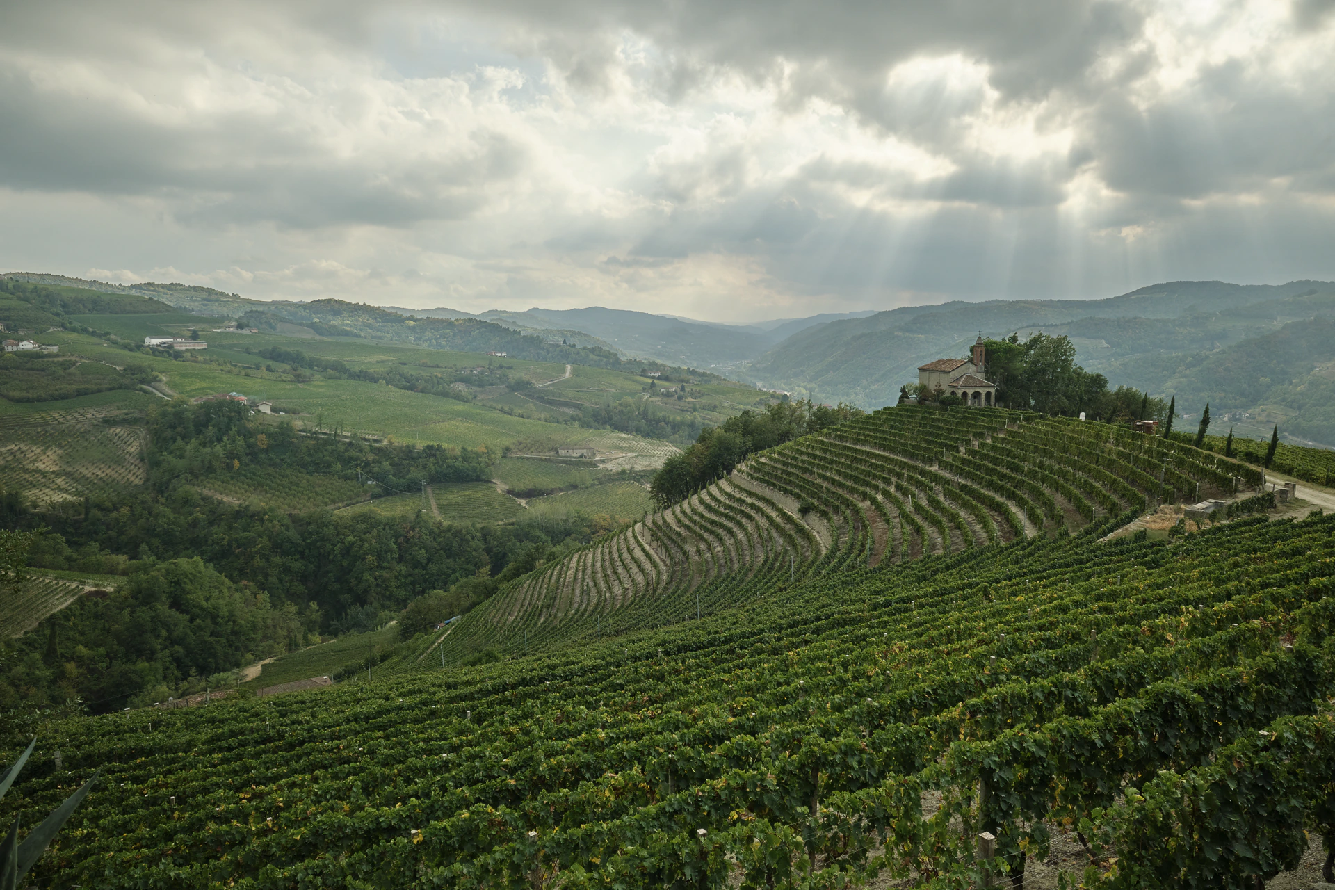 Aerial view of a hilltop chapel above the terraced vineyards of Vallebelbo, with sunbeams breaking through clouds