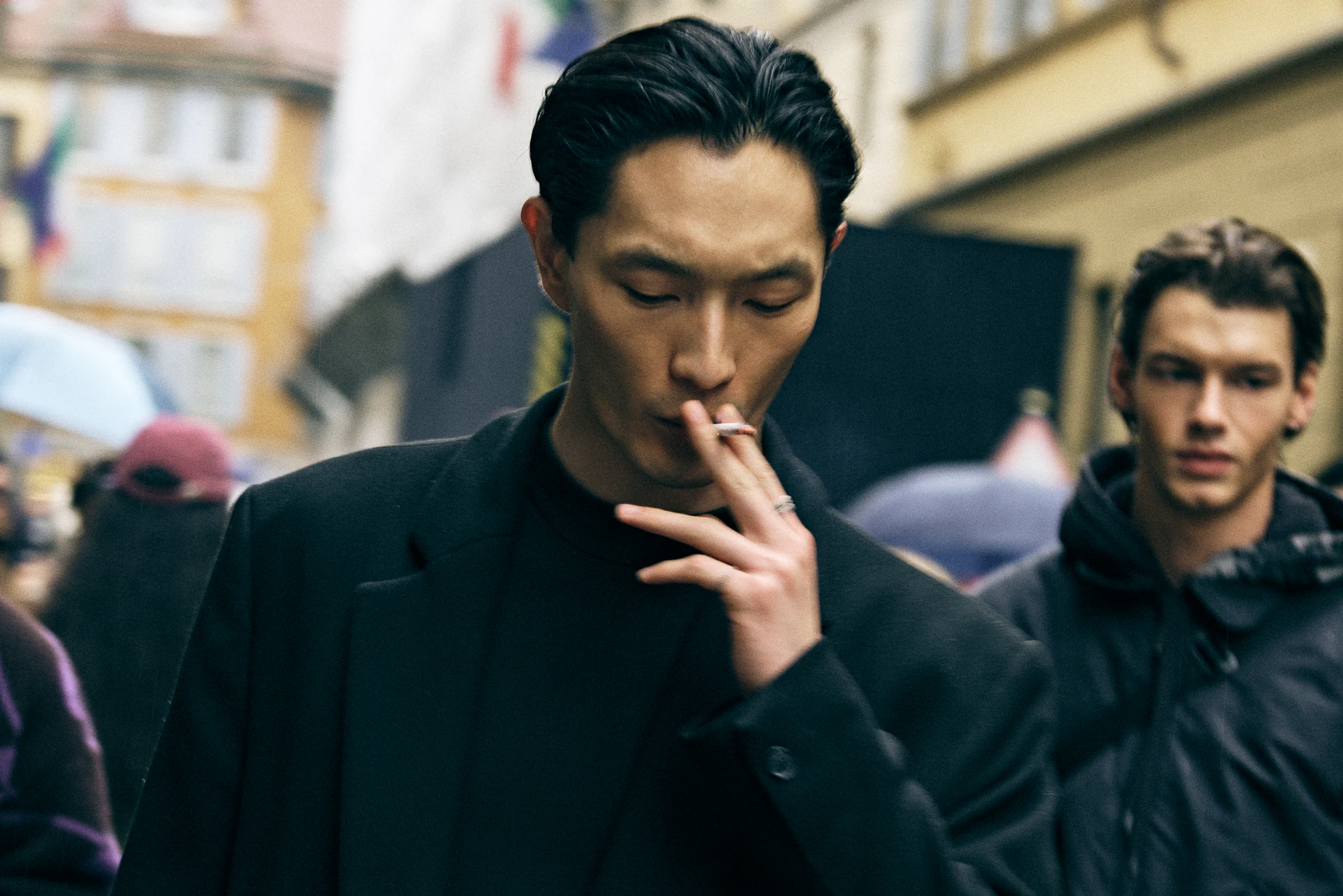 Close-up street style portrait of a man smoking in a dark coat, crowd blurred behind him at Milano Fashion Week