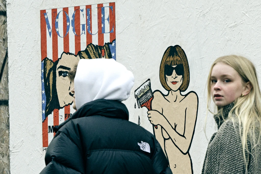 Two people passing a street art mural of Vogue magazine and Anna Wintour on a Milan wall, one wearing a black North Face puffer and white balaclava