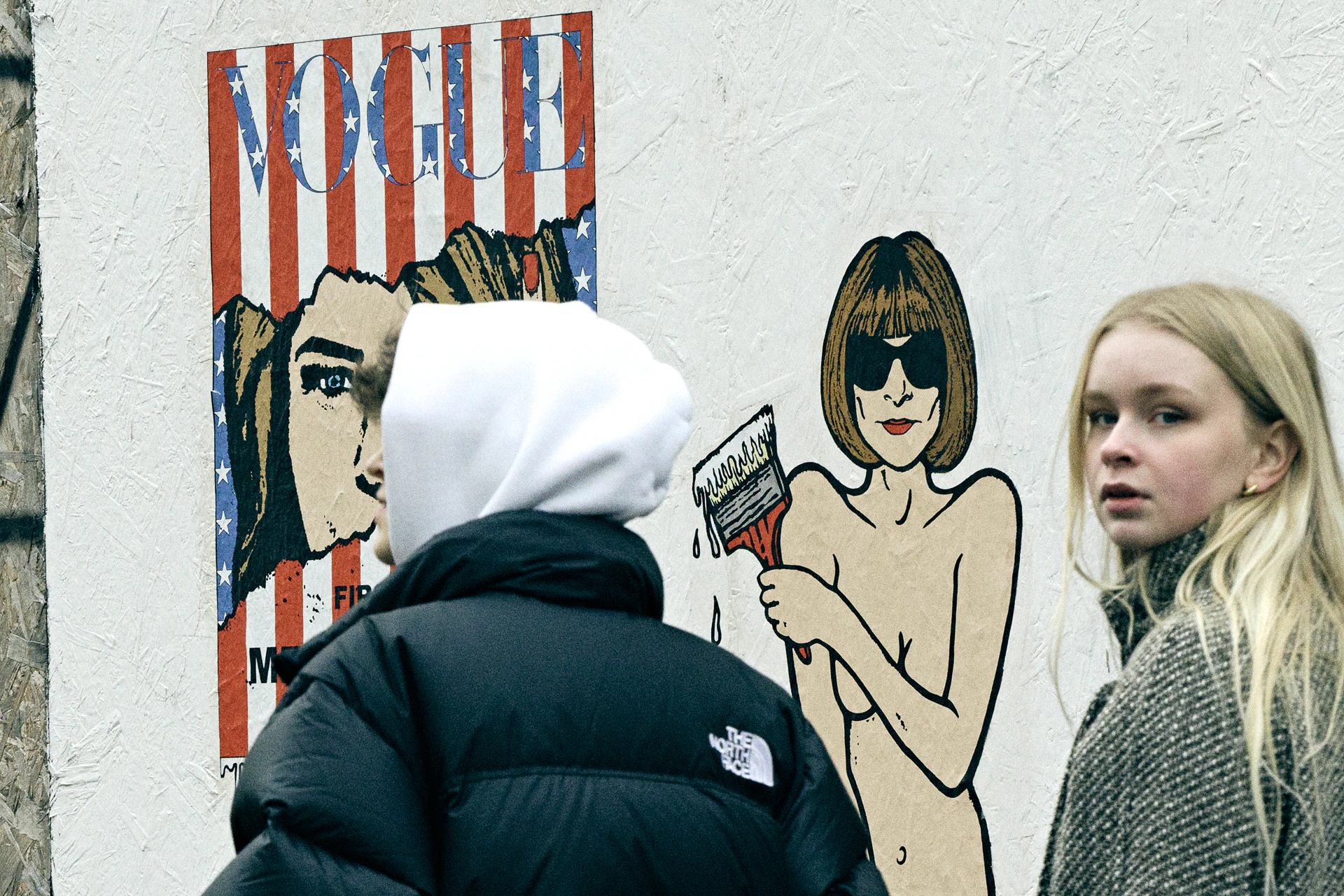 Two people passing a street art mural of Vogue magazine and Anna Wintour on a Milan wall, one wearing a black North Face puffer and white balaclava