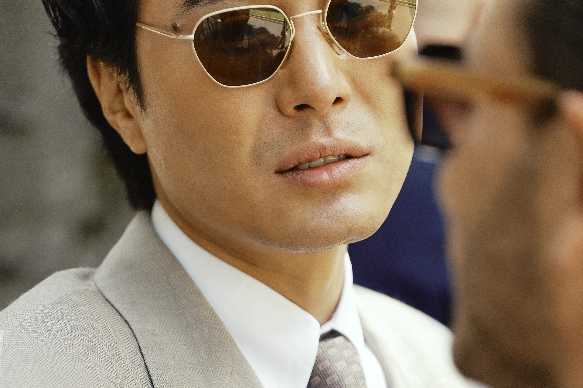 Close-up portrait of a man in a beige suit and tie wearing round gold-rim sunglasses with brown lenses, Milano Fashion Week street style