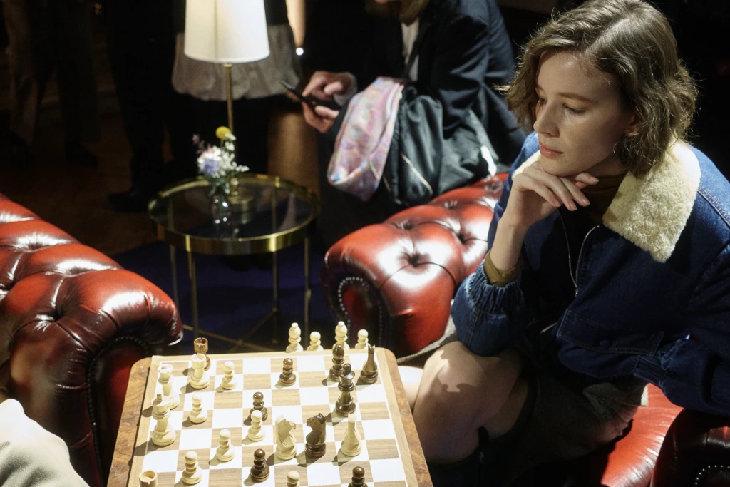 Woman resting her chin on her hand while watching a chess game on a red leather couch, Milano Fashion Week, January '26