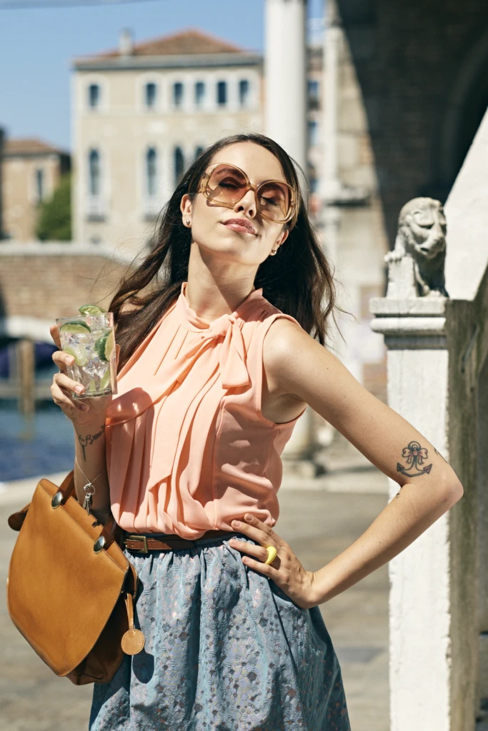Model in a peach blouse holding a citrus cocktail near a Venice canal, lion statue in the background