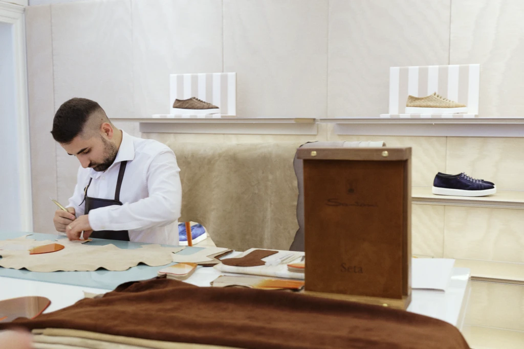 Craftsman at a Santoni leather workbench cutting shoe patterns, sneakers displayed on shelves behind him