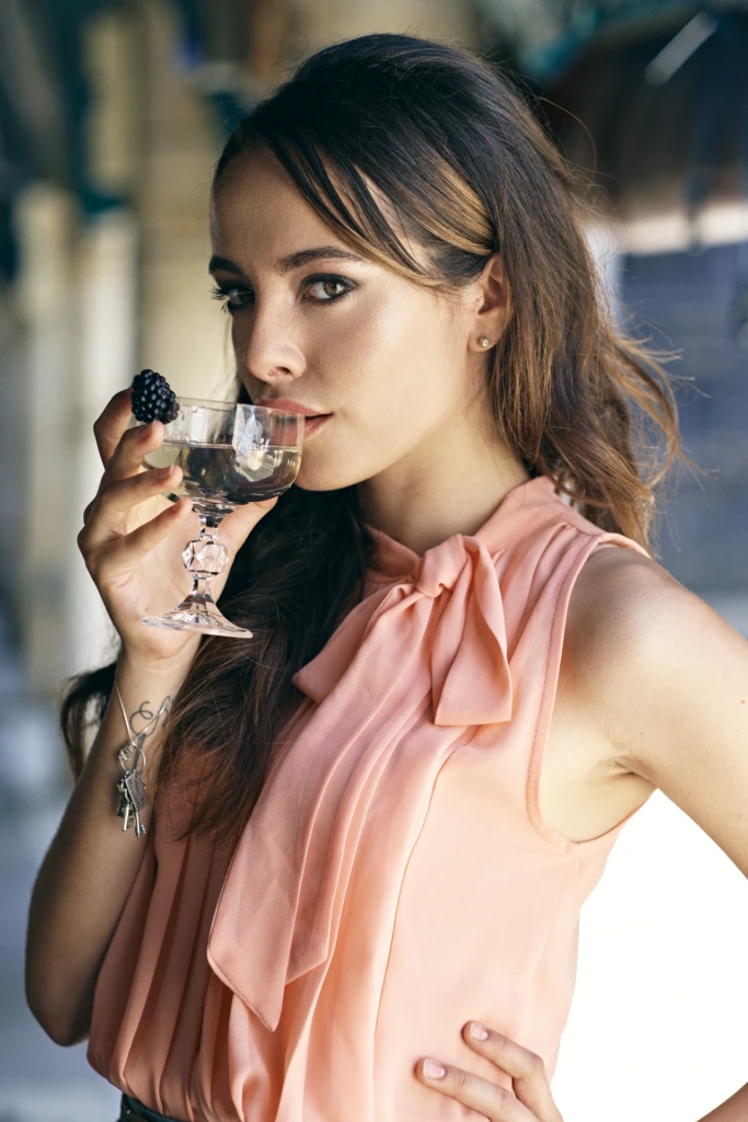 Close-up portrait of a model sipping a cocktail garnished with a blackberry, a key charm bracelet on her wrist