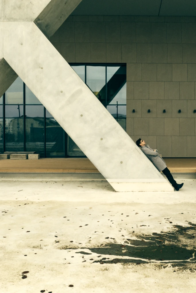 Marco Castelli in a grey coat leans against angular concrete architecture, looking up, at Milano Fashion Week
