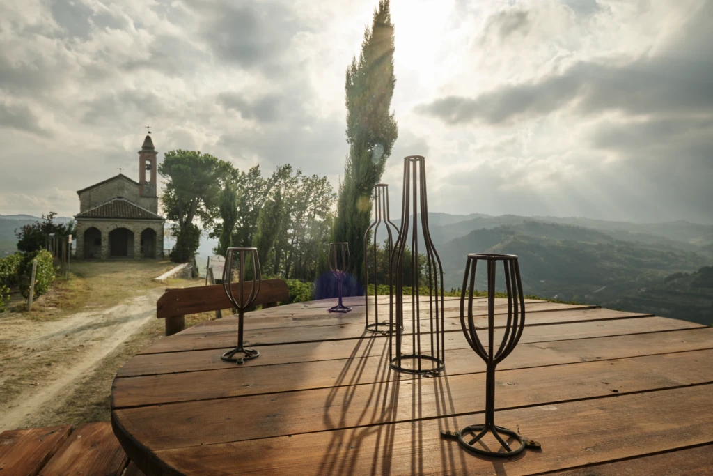 Iron wine bottle and glass sculptures on a wooden table overlooking the valley at golden hour, with the chapel in the distance