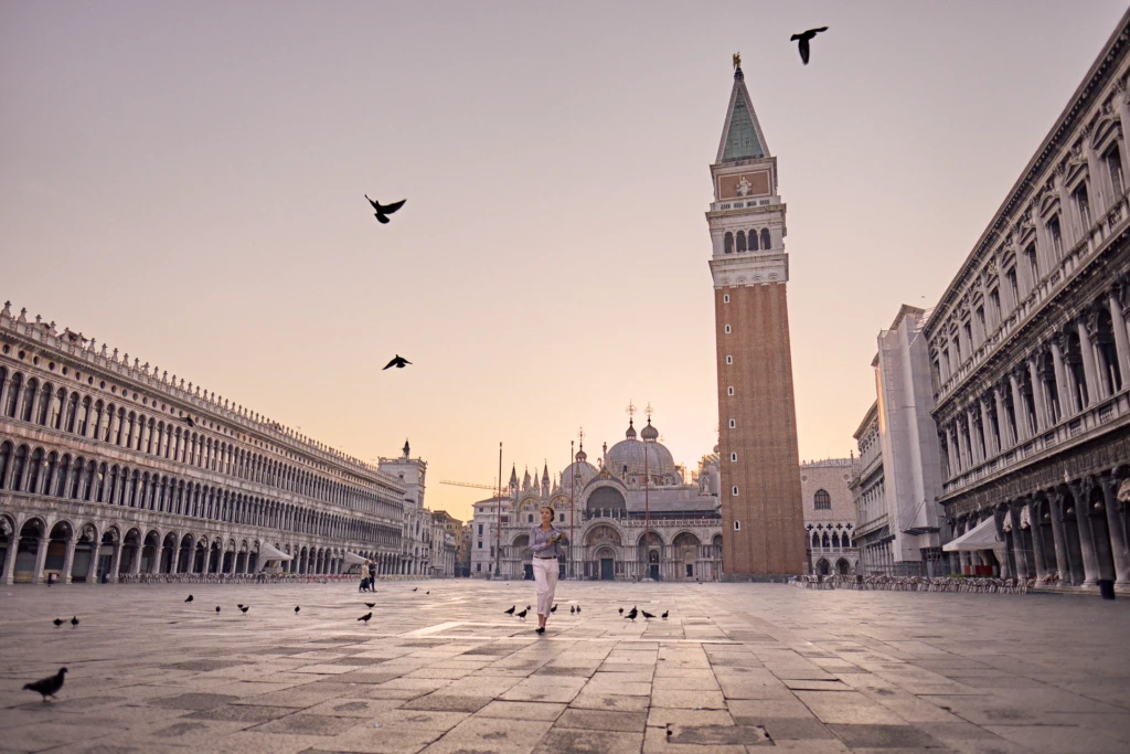 Wide view of a woman crossing Piazza San Marco at sunrise, the campanile and basilica behind her and pigeons in flight