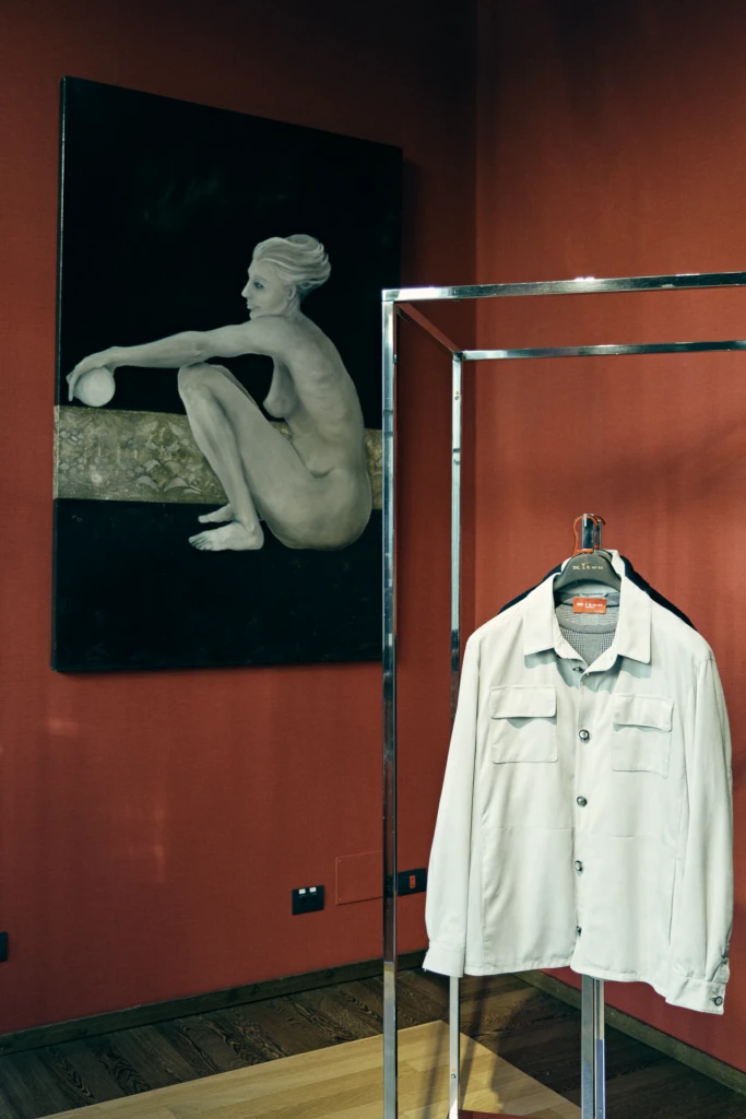 A dark surrealist nude painting beside a beige Kiton jacket displayed on a chrome rack in a red showroom