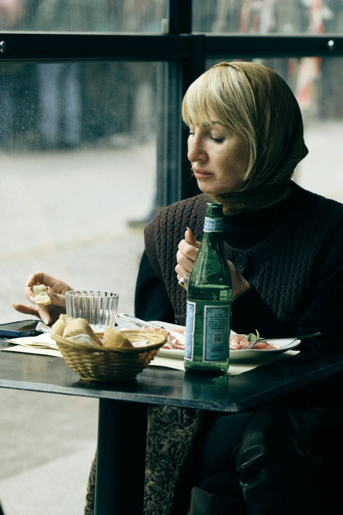 Woman in a headscarf eating bread at a cafe table with a bottle of sparkling water, Milano Fashion Week, January '26