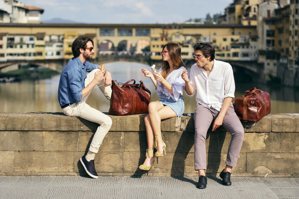 Couple walking hand in hand toward a stone archway gate in a fortified Tuscan hill town, grey Fiat 500 parked nearby, tan leather bag in hand