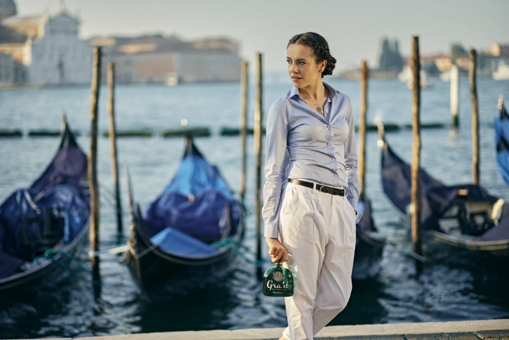 Model in a light blue shirt walking past Venice's gondolas, holding a bottle of Gra'it grappa
