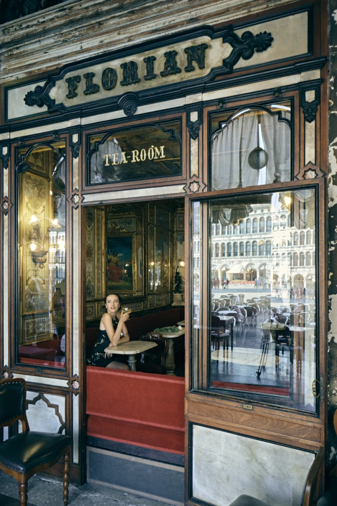 Model seated inside Caffè Florian's Tea Room with a cocktail, Piazza San Marco reflected in the storefront windows