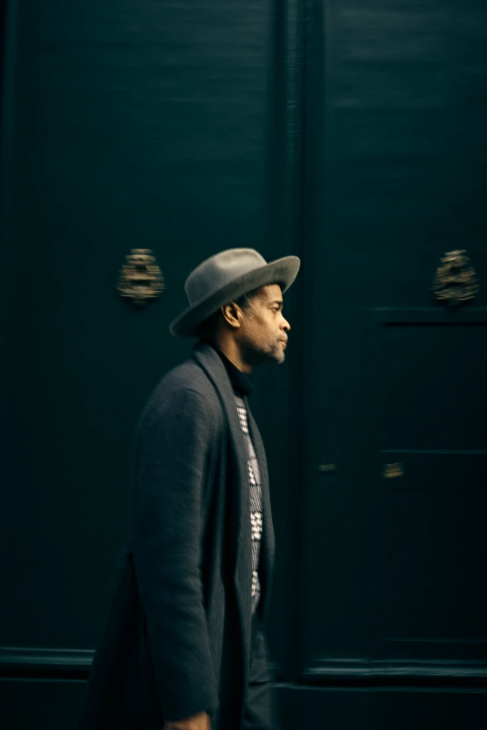Guerre, street style portrait in a dark coat and grey fedora against a dark green door in Milan