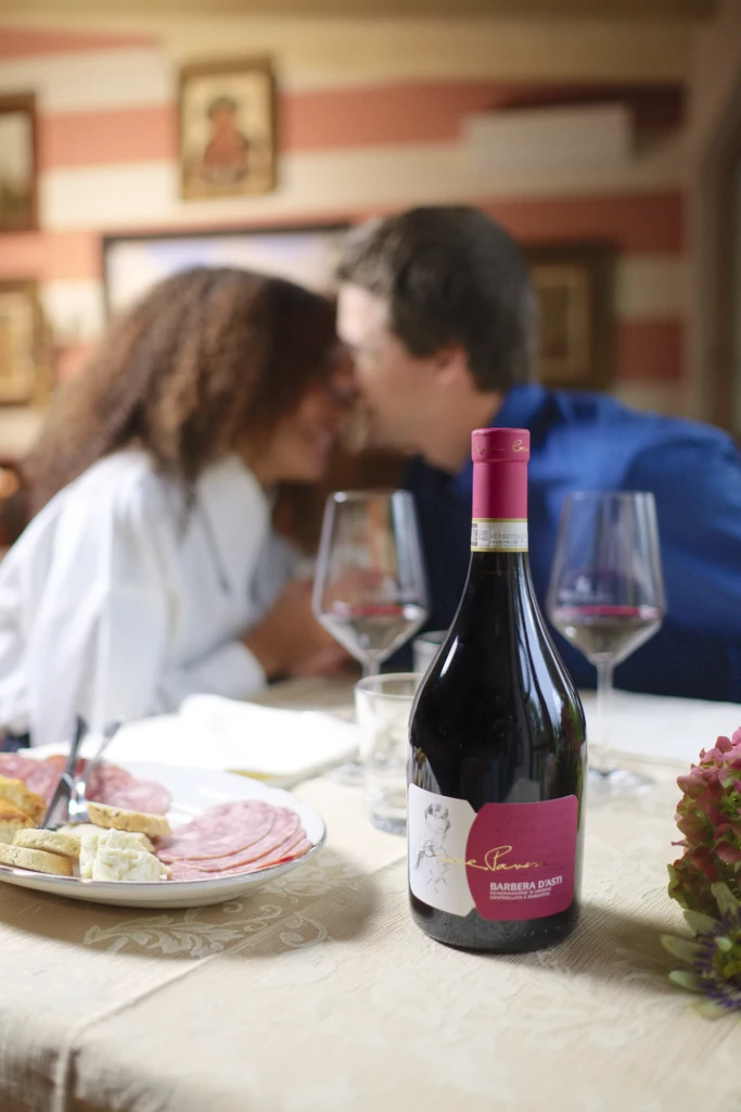 Two models embracing at the table with a bottle of Barbera d'Asti and wine glasses nearby