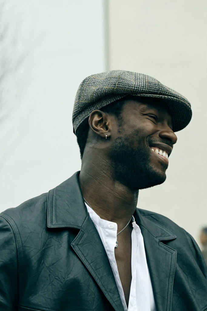 Smiling man in a checked flat cap and dark leather jacket over a white shirt, Milano Fashion Week street style, January '26