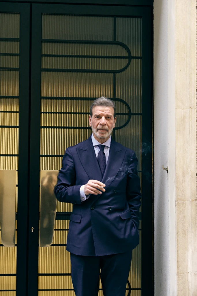 Street style portrait of a silver-haired man in a navy suit smoking a cigarette outside a gold-paneled door