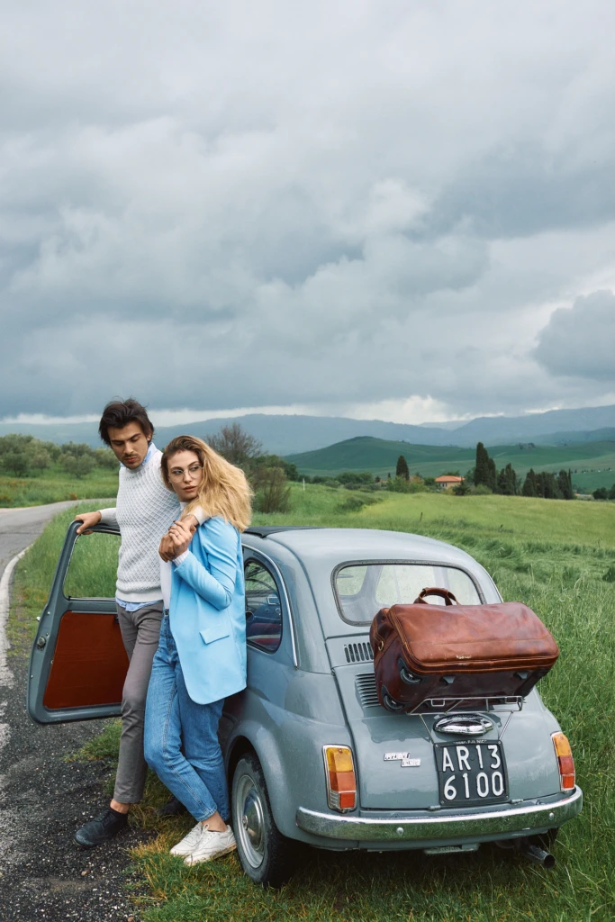 Couple leaning against a vintage grey Fiat 500 on a rain-soaked country road, tan leather duffel bag on the roof rack, storm clouds over Tuscan hills