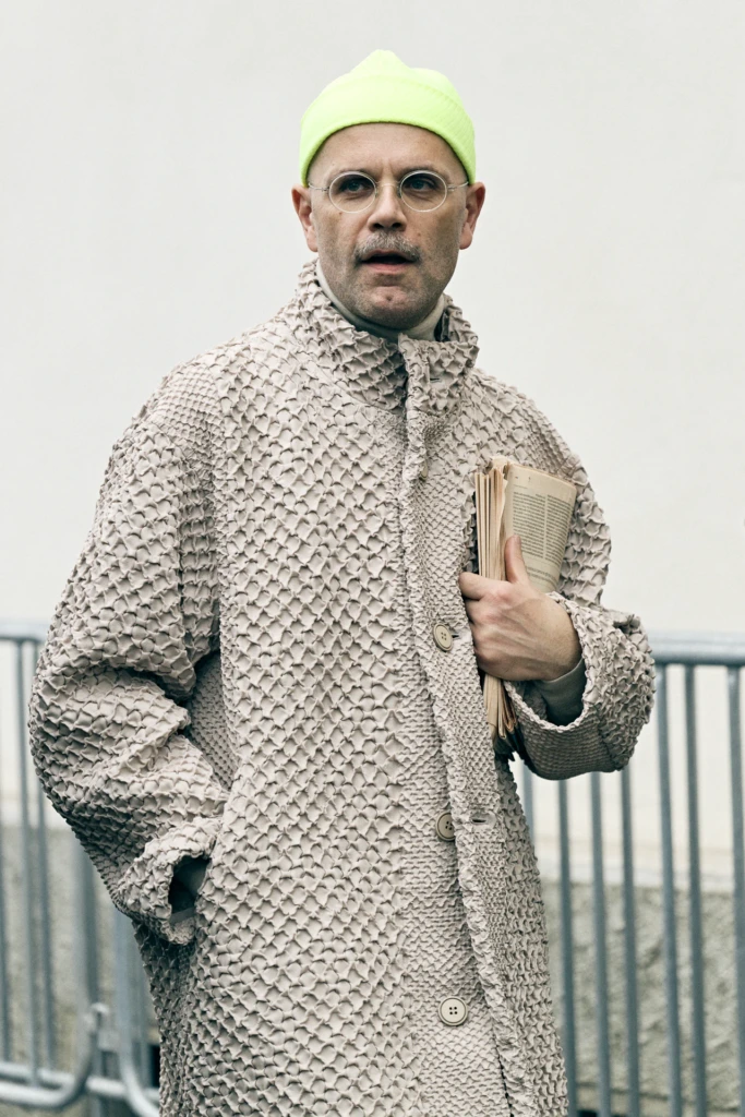 Man in a neon green beanie and round glasses carrying a folded newspaper, wearing a textured beige coat, Milano Fashion Week, January '26