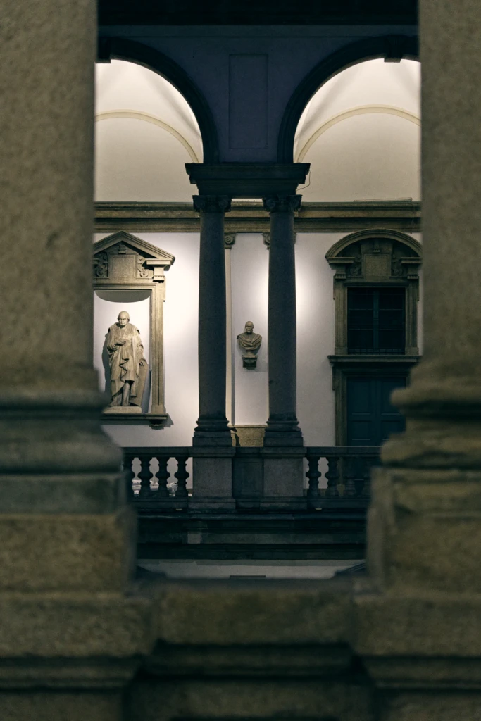 A neoclassical courtyard glimpsed through columns, with a statue and bust set into the walls