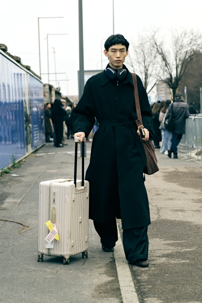 Young man in a long black trench coat pulling a white Rimowa suitcase down a Milan street, headphones around his neck, Milano Fashion Week, January '26