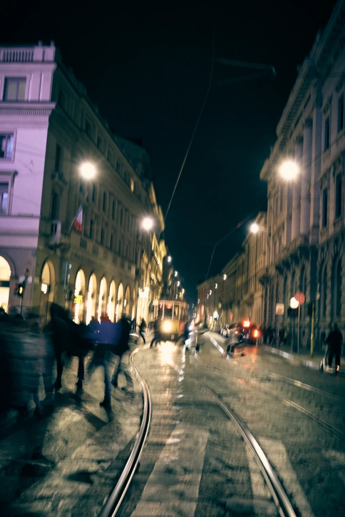 A blurred night street scene in Milan with a passing tram and pedestrians crossing the tracks