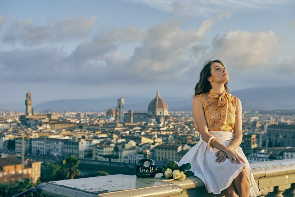 Model in a paisley blouse and white skirt gazing over the Florence skyline with the Gra'it bottle and white roses on a rooftop balustrade