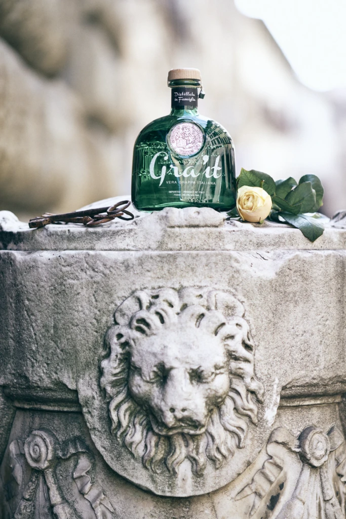 Gra'it grappa bottle, an antique key, and a white rose staged on a carved stone lion-head fountain in Florence