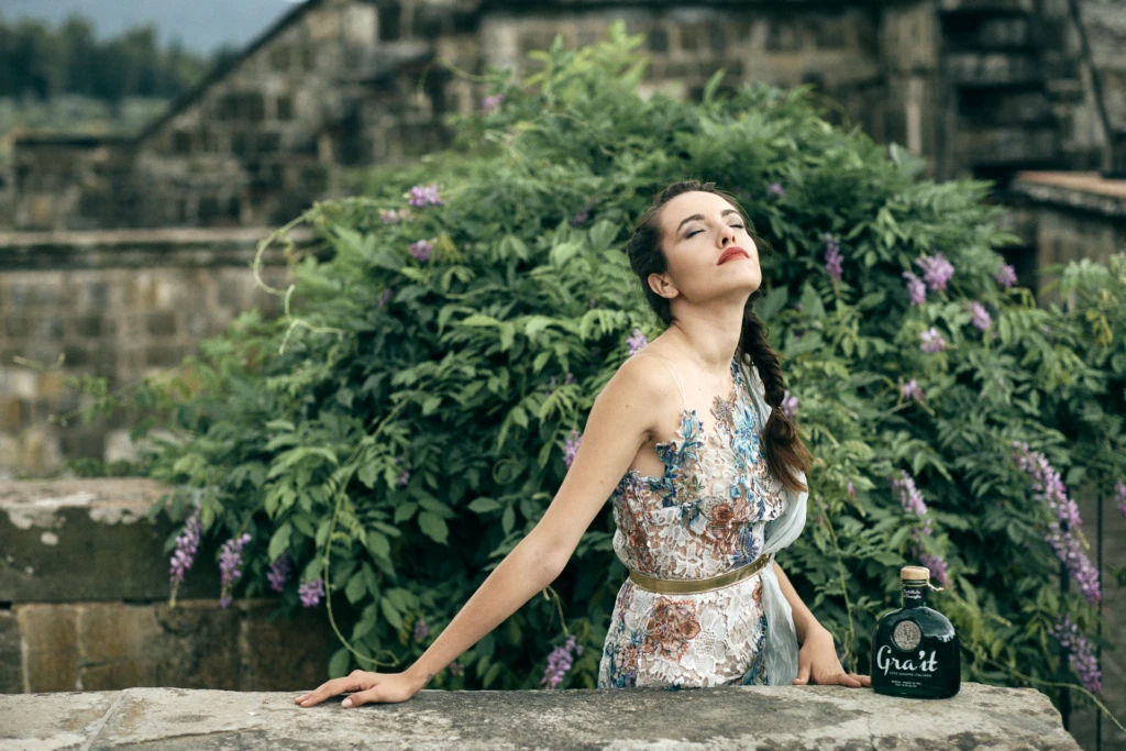 Model in a floral lace dress leaning against a wisteria-covered stone balustrade with the Gra'it bottle at a hillside villa