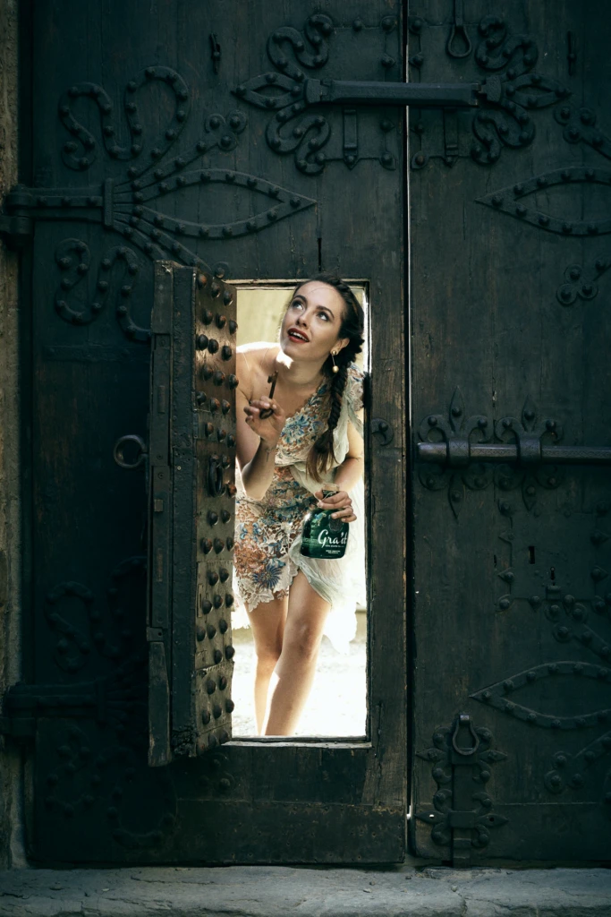 Model peering through an ornate wooden castle door, holding the Gra'it bottle and an antique key