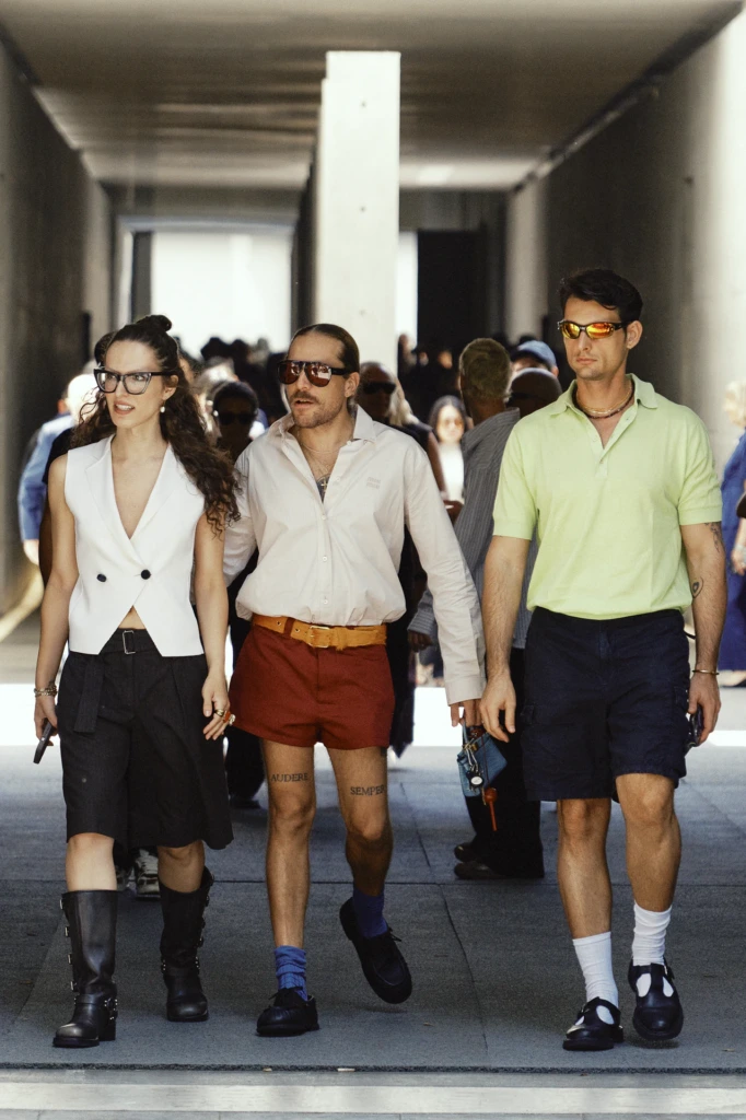 Three friends walking together in colorful Fashion Week outfits and sunglasses through a concrete venue corridor