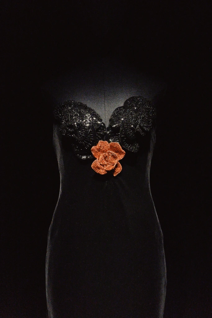 Black dress detail with sequined floral appliqué and an orange rose brooch, fashion exhibit lighting